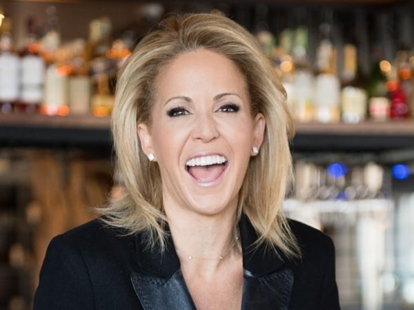 Smiling person in a black blazer in a blurred bar setting, with bottles on shelves behind them.