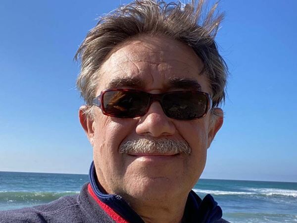 Man wearing sunglasses smiles at the beach with ocean and blue sky behind him