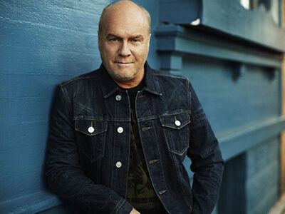 Man in a dark denim jacket leaning against a blue wall outdoors