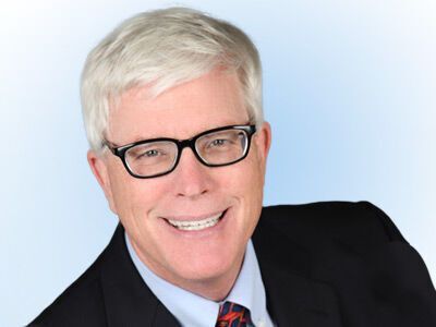 Smiling man with white hair and black glasses in a dark suit against a light blue background