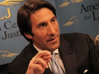 Man in dark suit speaking into a microphone at a conference, with blue backdrop and partial “Justice” text