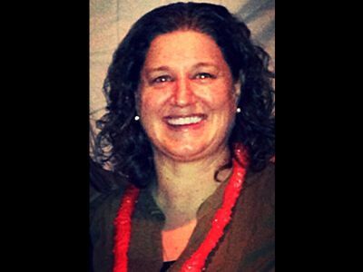 Smiling person with curly dark hair wearing a red necklace against a dark background