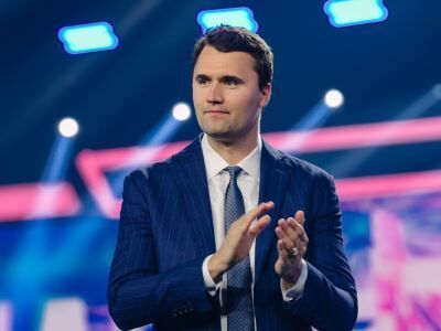 Man in a blue suit clapping on a brightly lit stage with blurred neon lights behind him