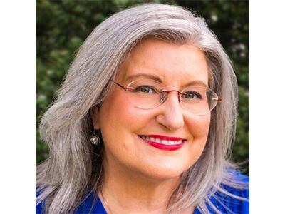 Smiling woman with gray hair, glasses, and red lipstick in a blue top against a blurred outdoor background