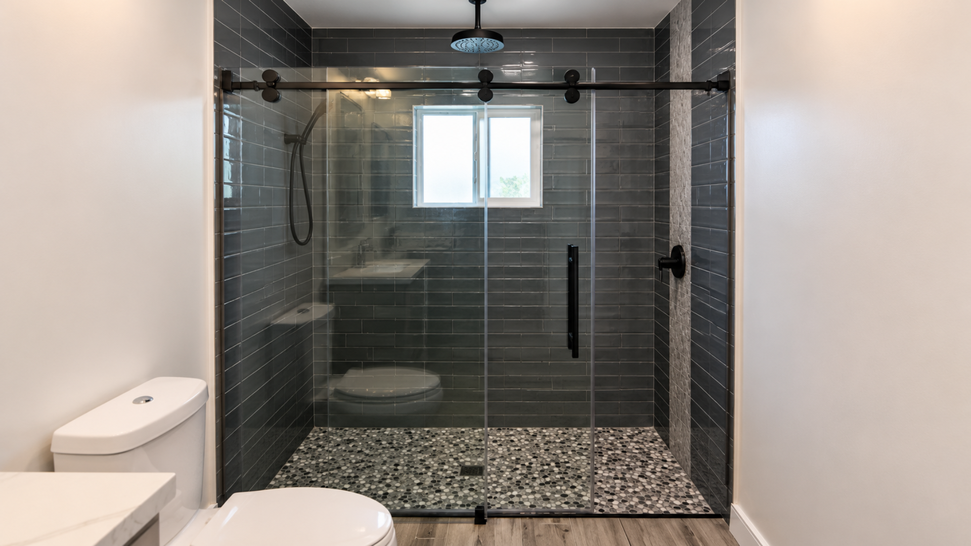 Modern bathroom with glass-enclosed shower, dark tile walls, pebble floor, and a small window.