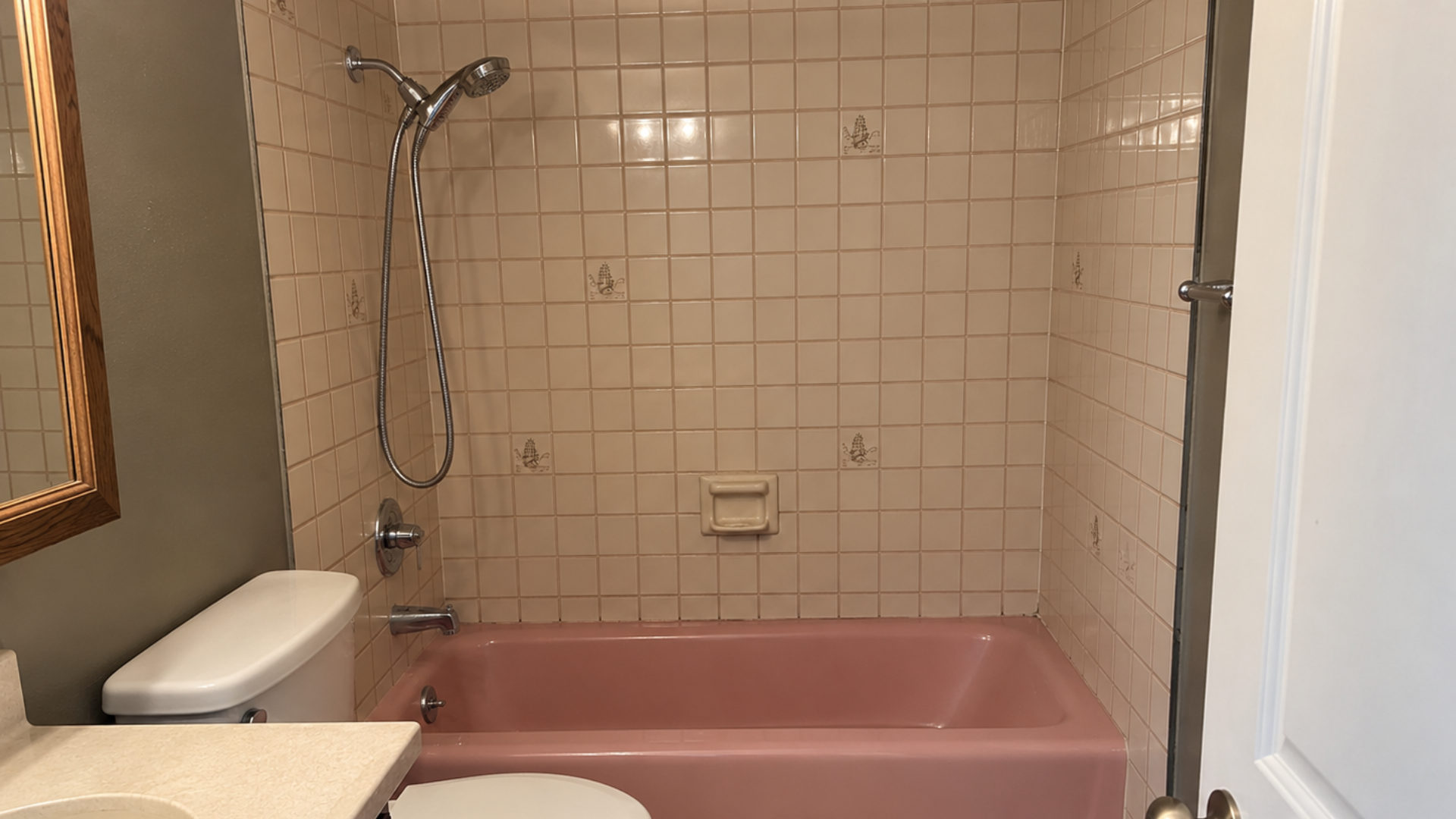 Small bathroom with pink bathtub, white tiled walls, toilet, and showerhead under a window.