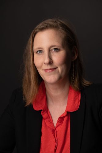 Dorit Horst, bras croisés, vêtue d'un blazer et d'une chemise rayée, souriante. Fond gris.