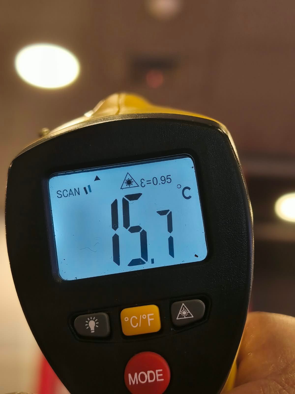 Termómetro digital que muestra 15,7 °C en una pantalla iluminada en azul, sostenido en la mano en el interior de una vivienda.