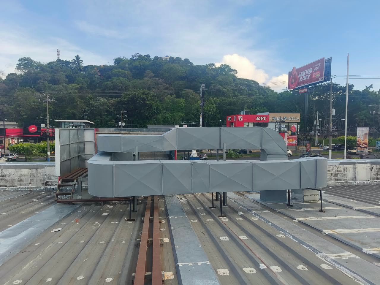 Unidades de climatización en la azotea de un edificio industrial plano, con montañas y vallas publicitarias al fondo.