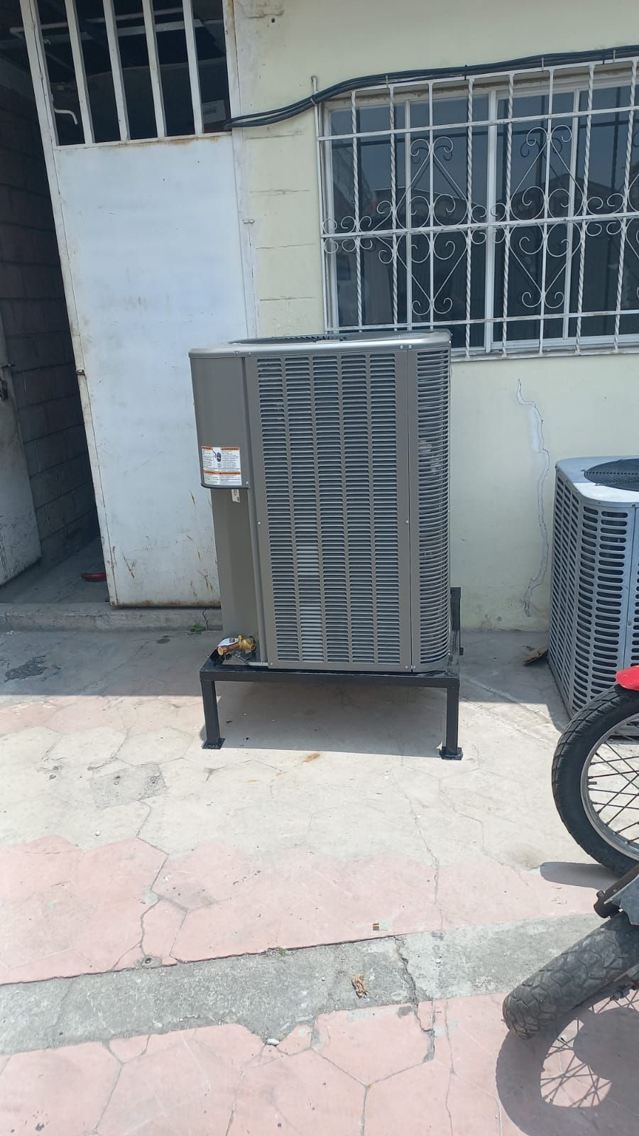Unidad exterior de climatización situada frente a un edificio, con ventanas enrejadas y parte de la rueda de una motocicleta visible.