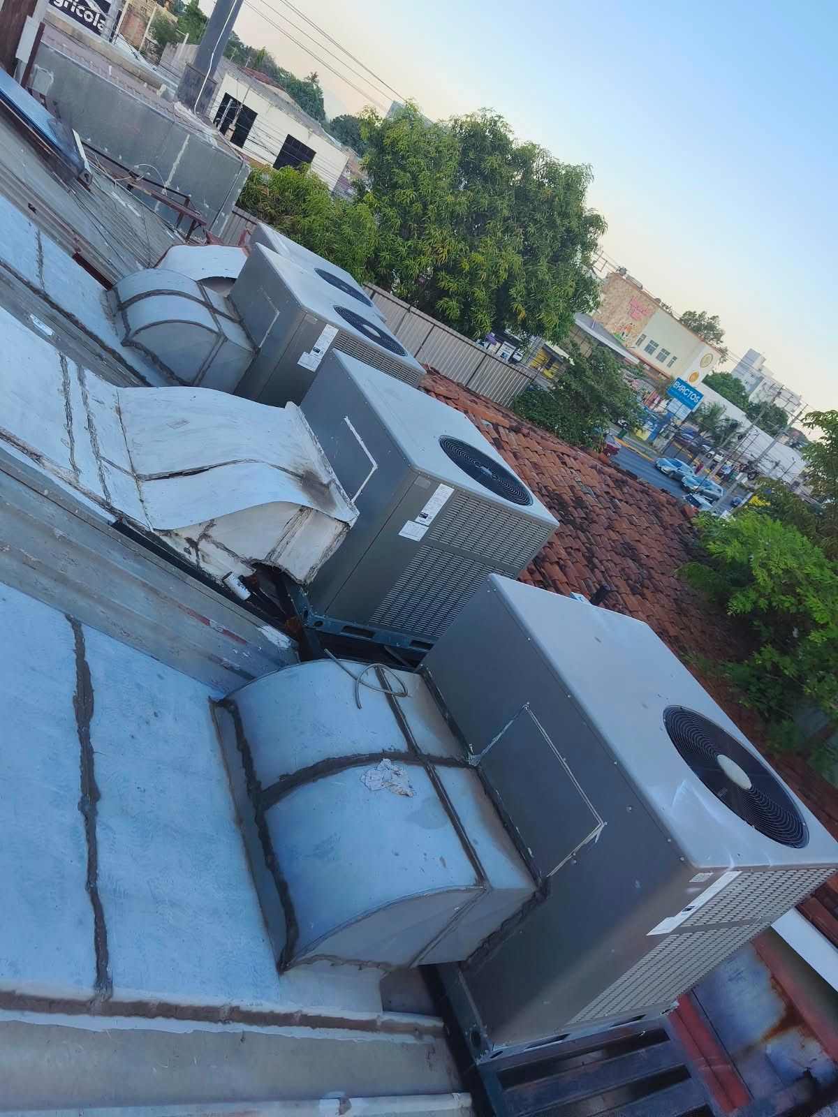 Unidades de aire acondicionado y rejillas de ventilación en la azotea, junto a una pared de ladrillos y árboles verdes al anochecer.