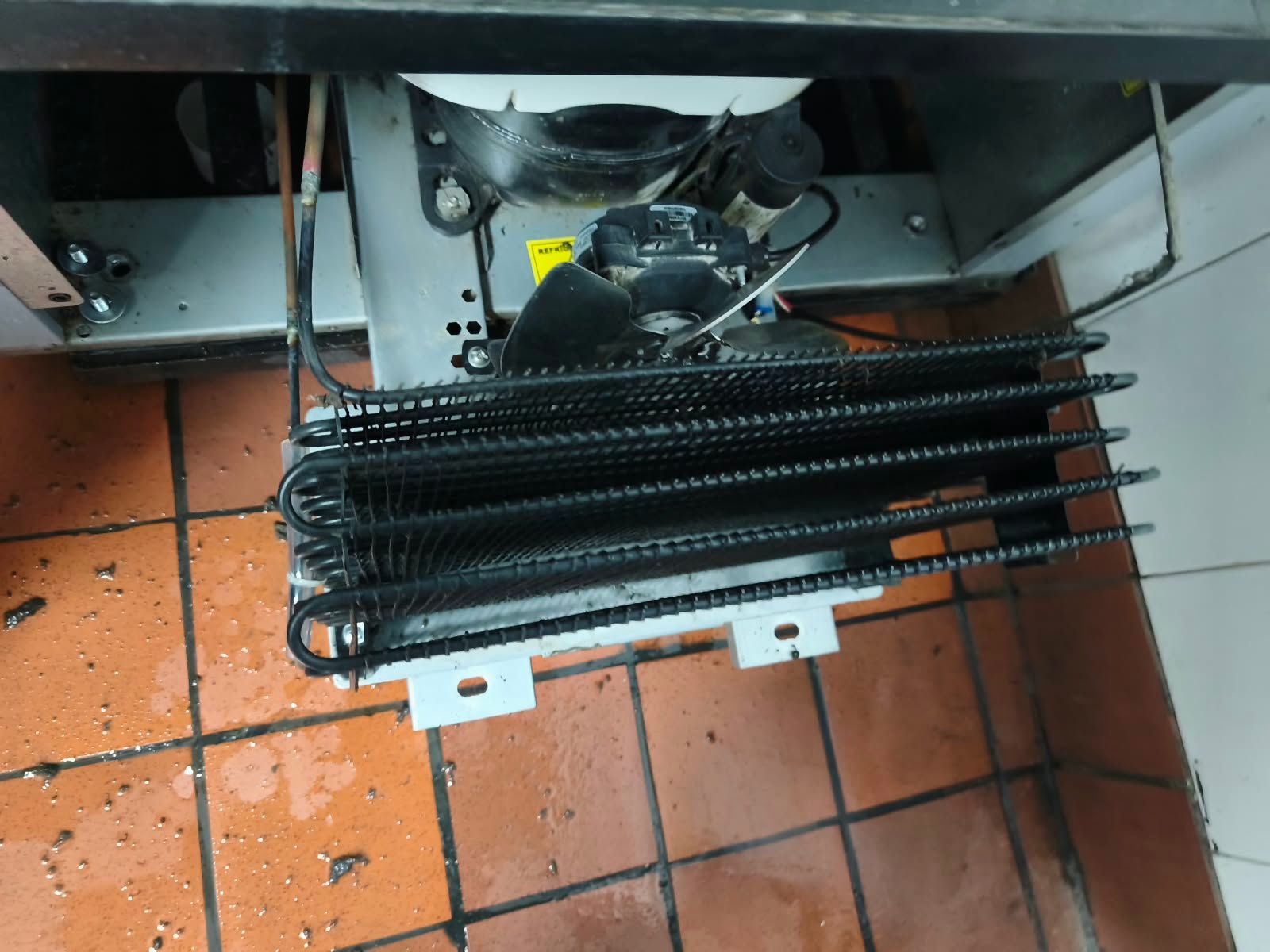 Serpentín de refrigeración de metal negro debajo de la maquinaria sobre un suelo de baldosas con escombros dispersos.