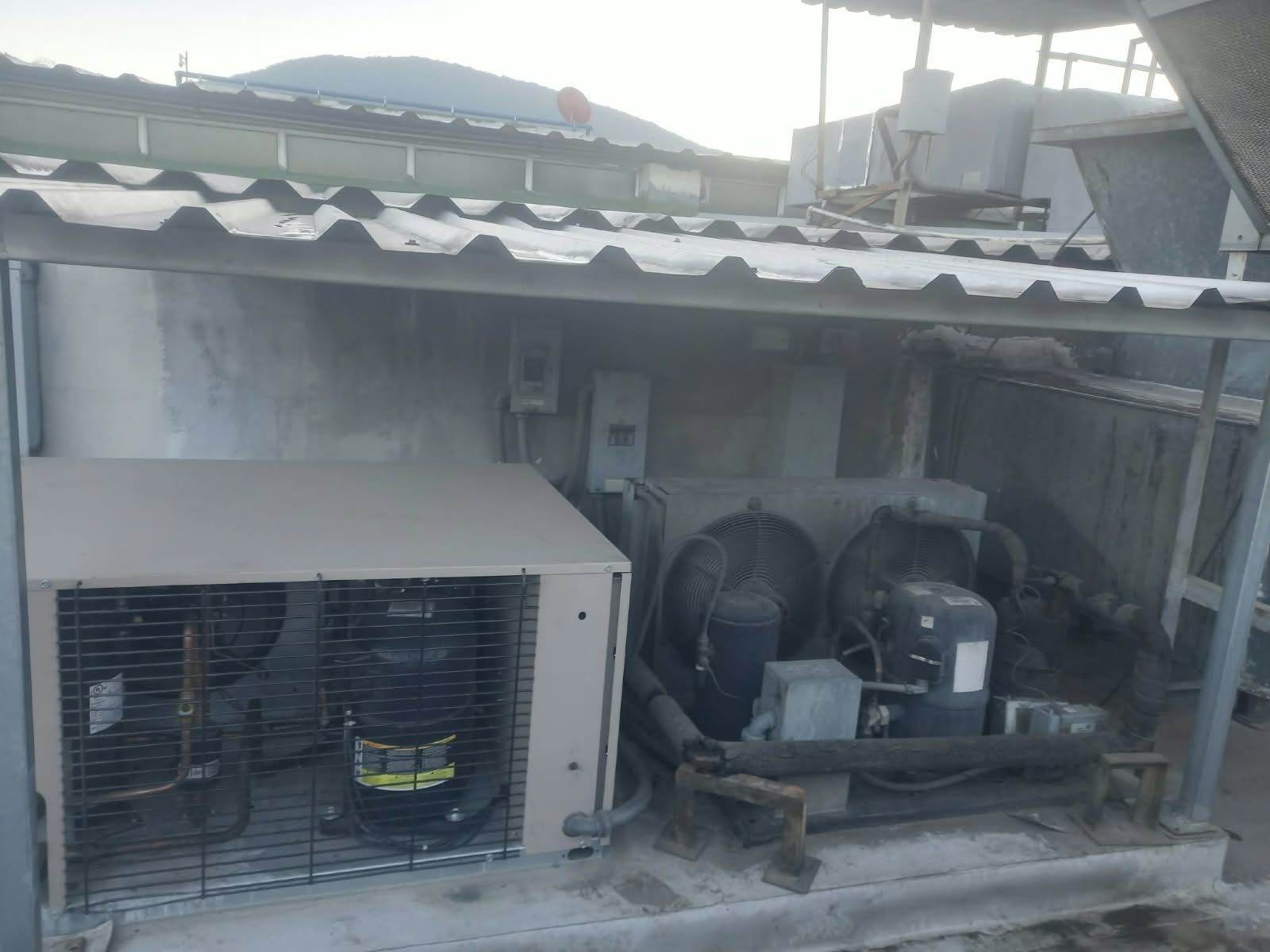 Unidades exteriores de aire acondicionado y compresores bajo un techo ondulado junto a una pared de hormigón.