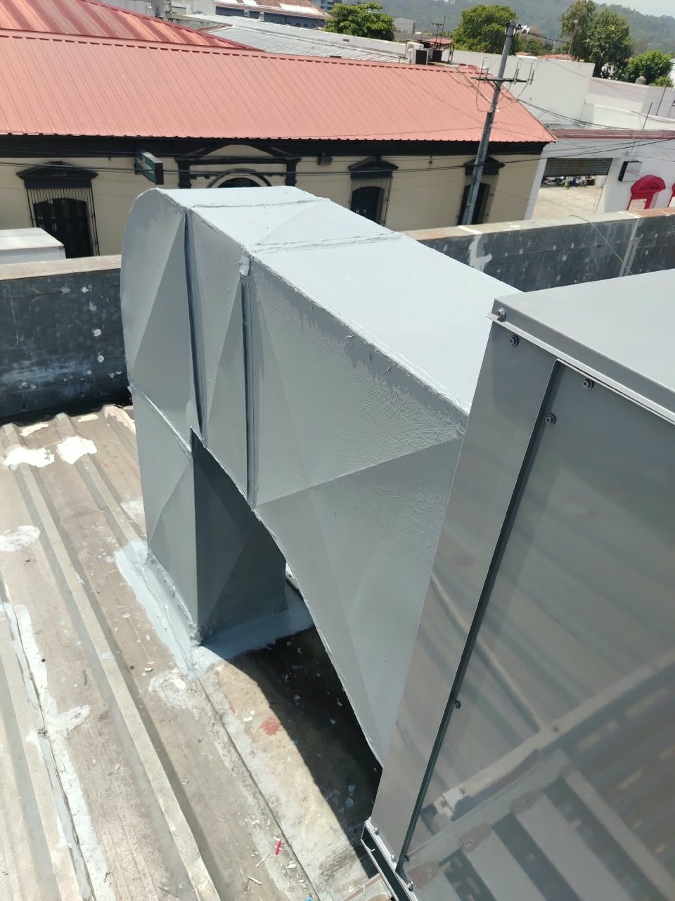 Conductos de climatización grises en la azotea de un tejado de chapa ondulada junto a un edificio de tejas rojas bajo la luz del día.