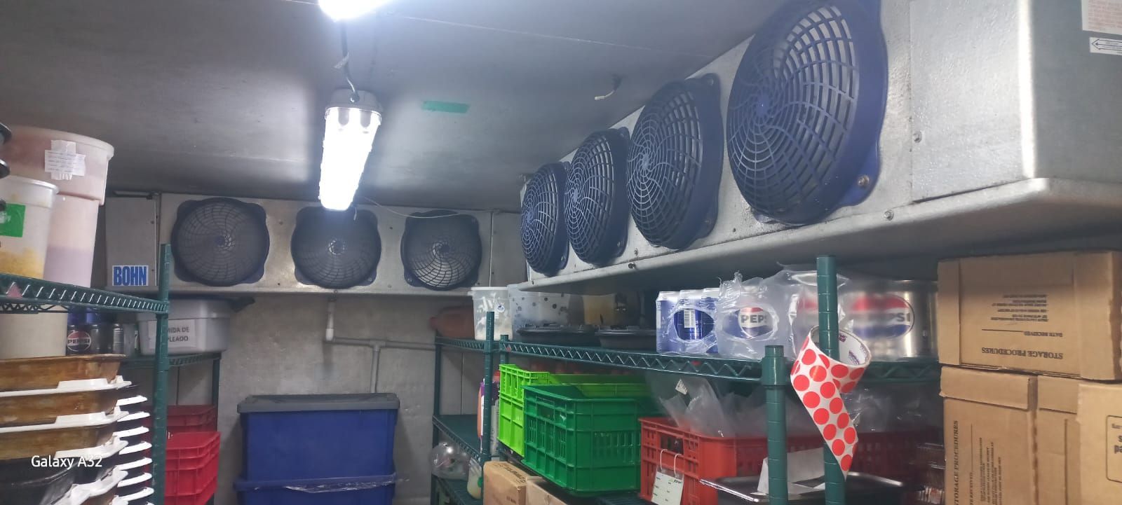 Trastero con estanterías llenas de cajas, contenedores de plástico y grandes ventiladores azules bajo luces fluorescentes.