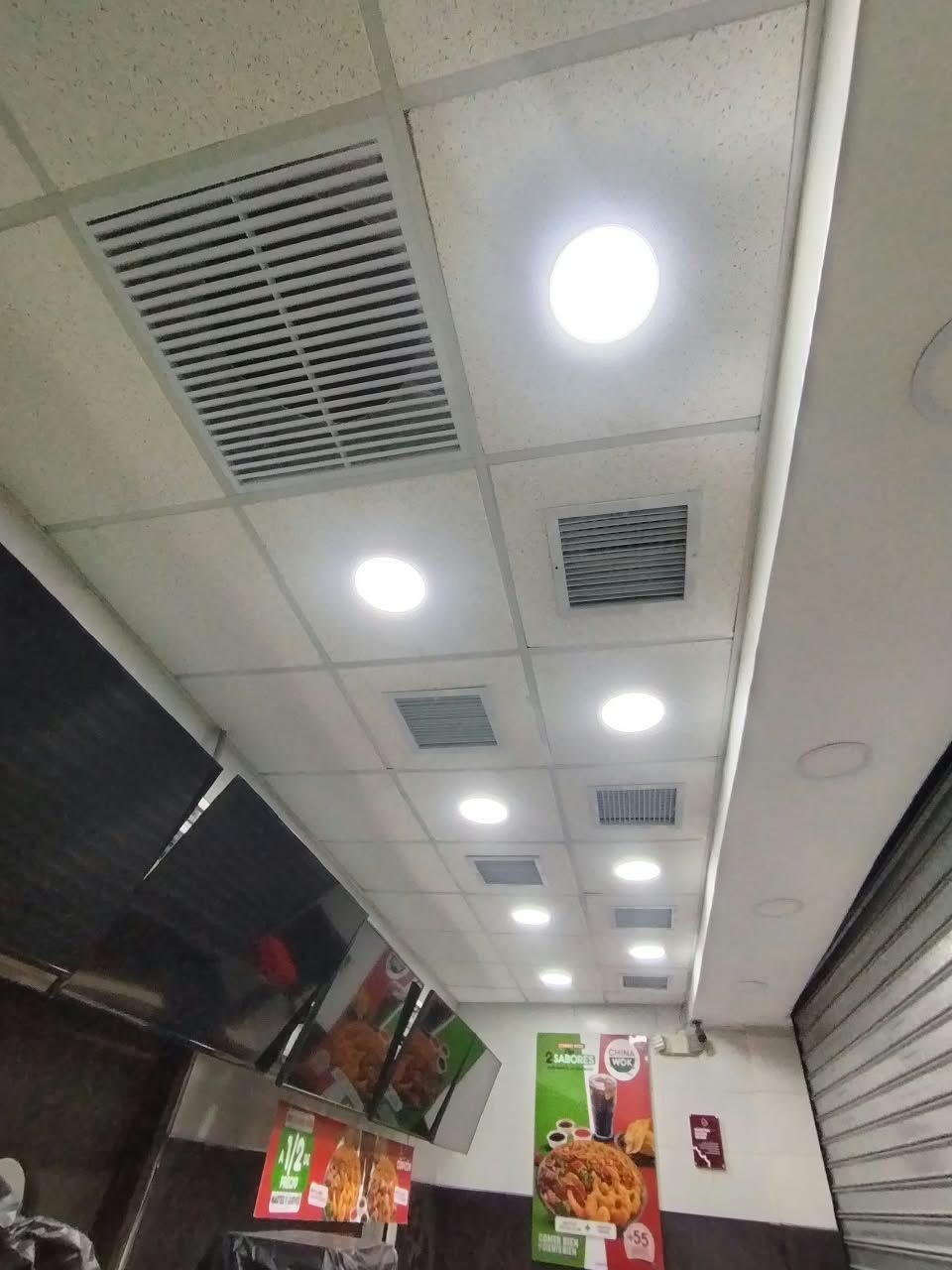 Vista inclinada del techo de un pasillo con luces empotradas, rejillas de ventilación y letreros colgantes de menús en una tienda o patio de comidas.