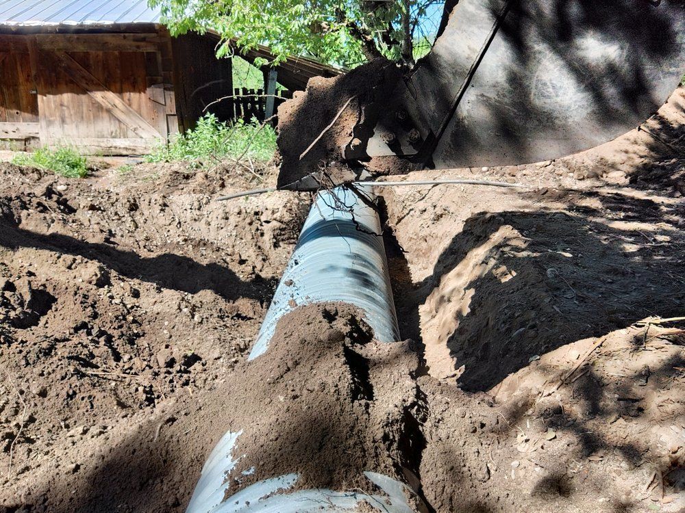 Large blue pipe laid in a dirt trench, with soil piled on both sides near a rural building.