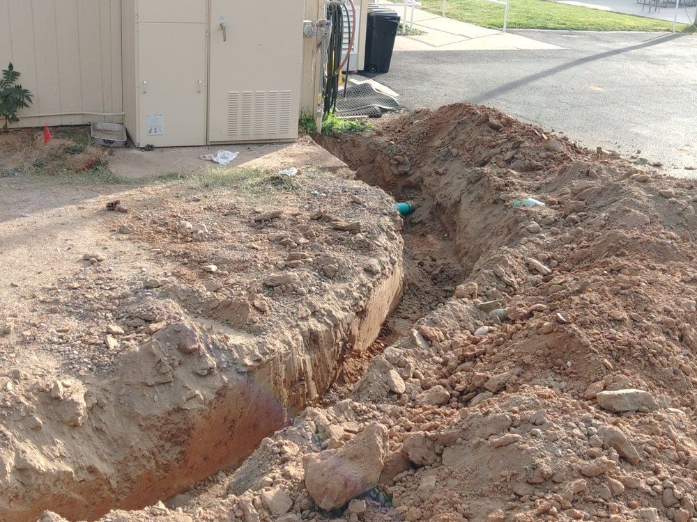 Large trench dug beside a building on a dirt lot, with piles of excavated soil along the edge.