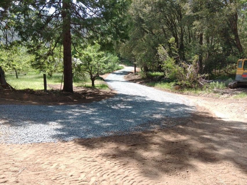 Gravel road winding through a wooded area, with sunlight and a parked vehicle at the right edge.