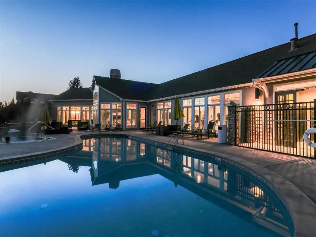 Outdoor swimming pool and hot tub area with lounge seating and a clubhouse in the background at dusk.