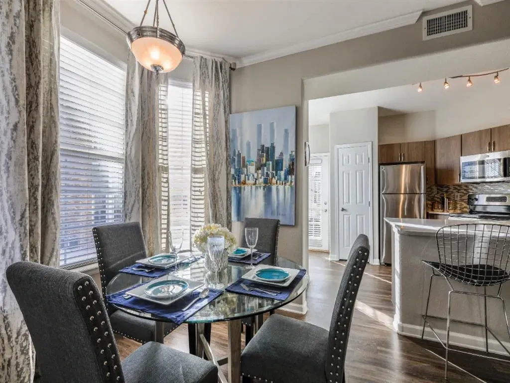 Dining area with a glass table and four chairs, next to a kitchen with stainless steel appliances.