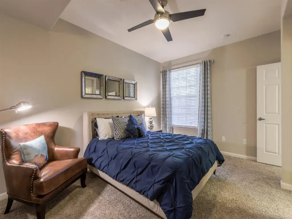Bedroom with a queen-size bed, dark blue comforter, two nightstands, a leather armchair, and a ceiling fan.