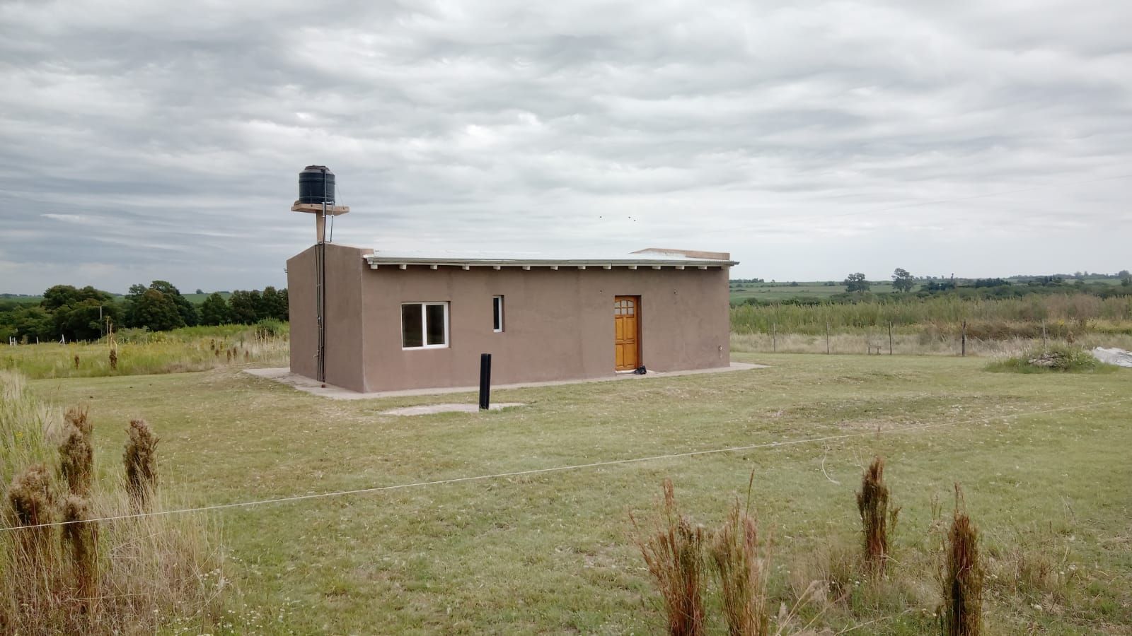 Pequeño edificio beige con un tanque de agua en un campo de hierba bajo un cielo nublado.