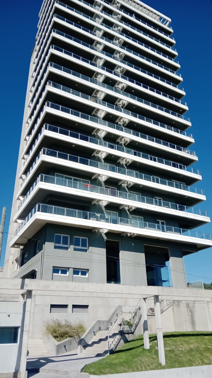 Moderno edificio de apartamentos de gran altura con balcones y una base escalonada de hormigón bajo un cielo azul despejado.