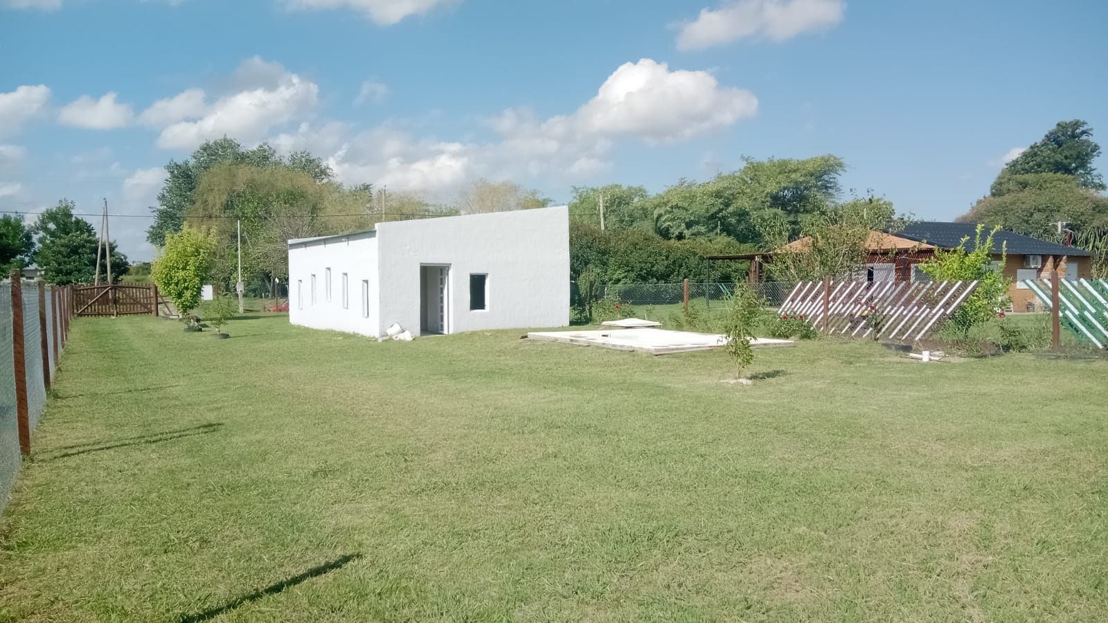 Sendero cubierto de hierba junto a edificios blancos y una valla de tela metálica bajo un cielo brillante.