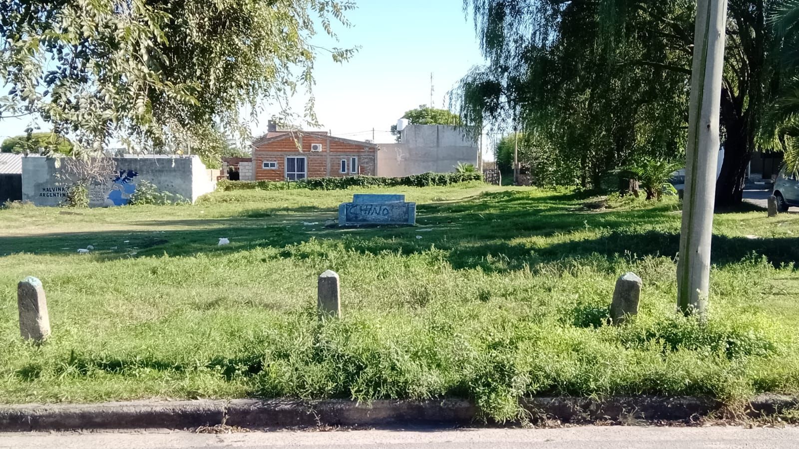 Un parque soleado y cubierto de césped, con árboles y una pequeña estructura blanca al fondo.