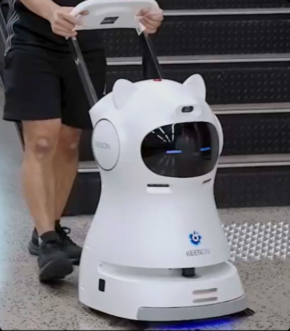 Person pushing a white, cat-faced cleaning robot with a Keelson logo. The robot is on a tiled floor.