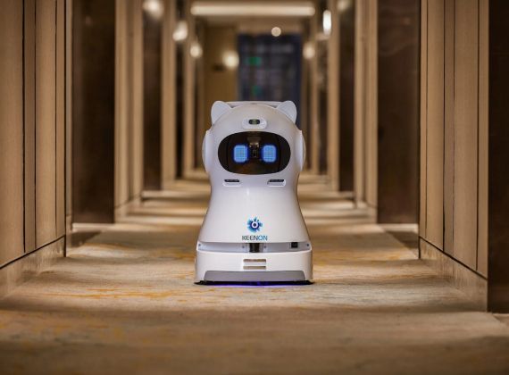 A white, cat-eared delivery robot with a digital blue-eyed face stands in the center of a hotel hallway.