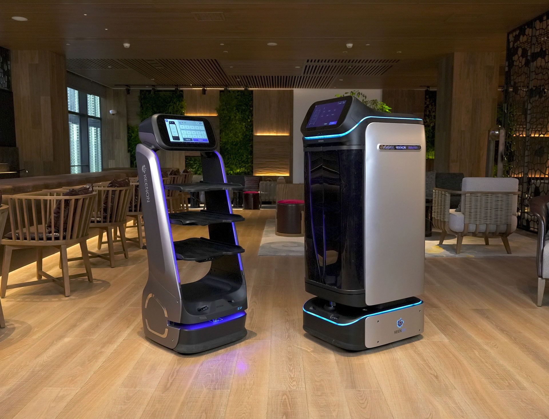 Two service robots in a restaurant setting. One has shelves, the other is taller with a screen.