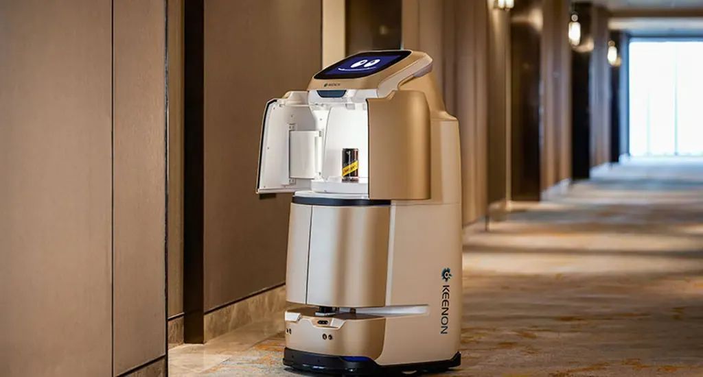 Hotel service robot in a hallway, beige body, carrying items, with a digital screen.