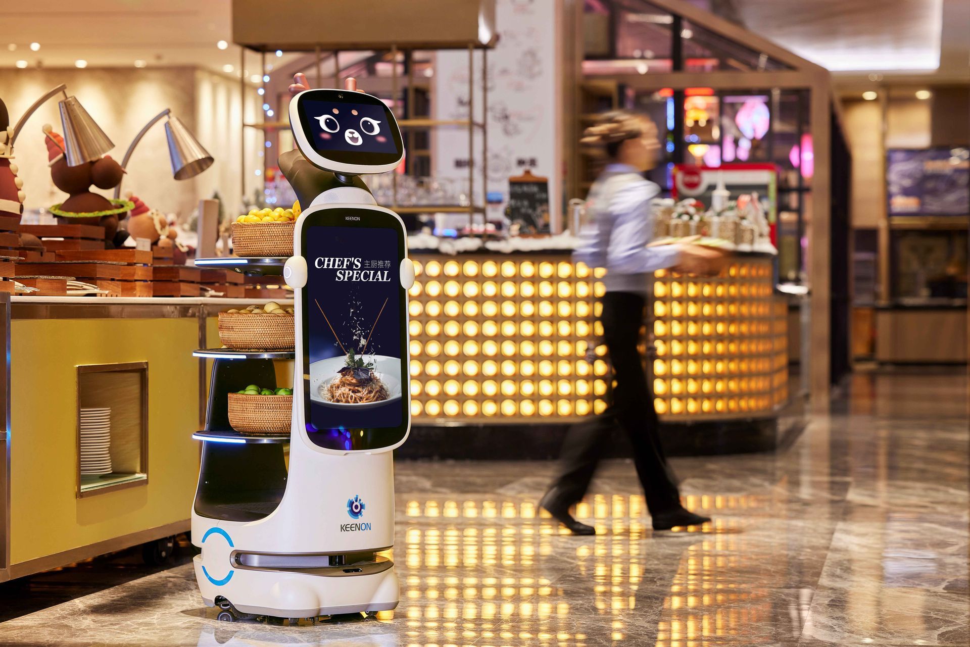 Robot server with food stands in front of a buffet, woman carries a tray in the background.