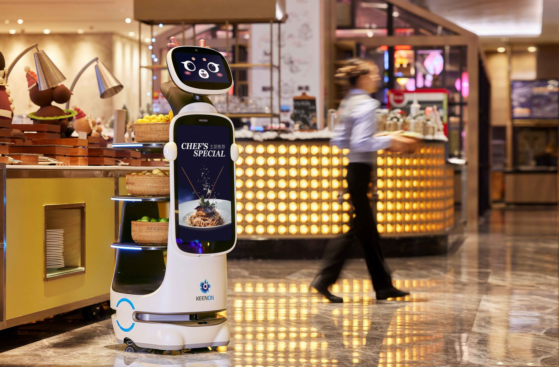A service robot with a digital screen face stands in a restaurant beside a person walking in the background.