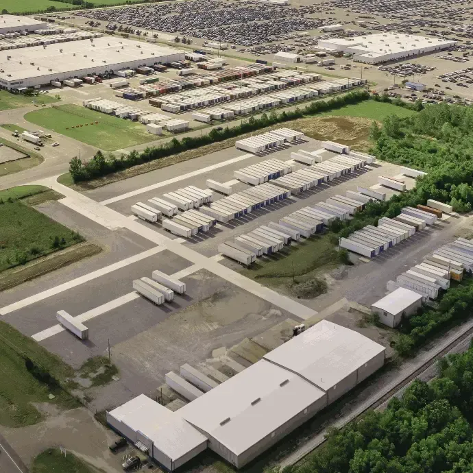 Aerial view of a large industrial warehouse yard with rows of white trailers and buildings near greenery.