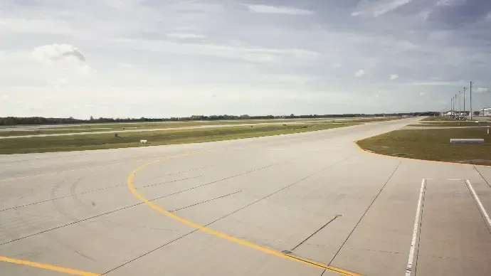 Empty airport runway curving right under a cloudy sky, with grassy fields and distant terminal lights.