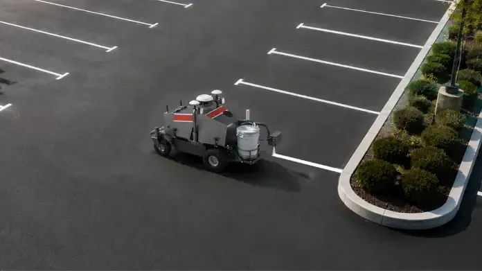 Small gray utility vehicle driving through an empty parking lot beside landscaped islands.