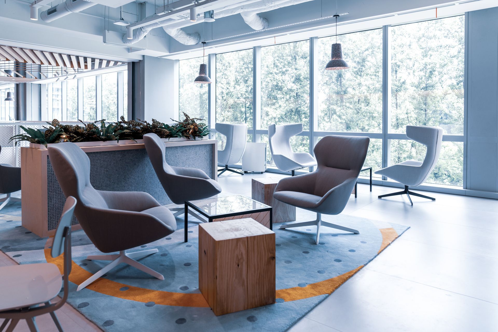 Modern office lounge with gray chairs, wooden tables, plants, and large windows overlooking trees