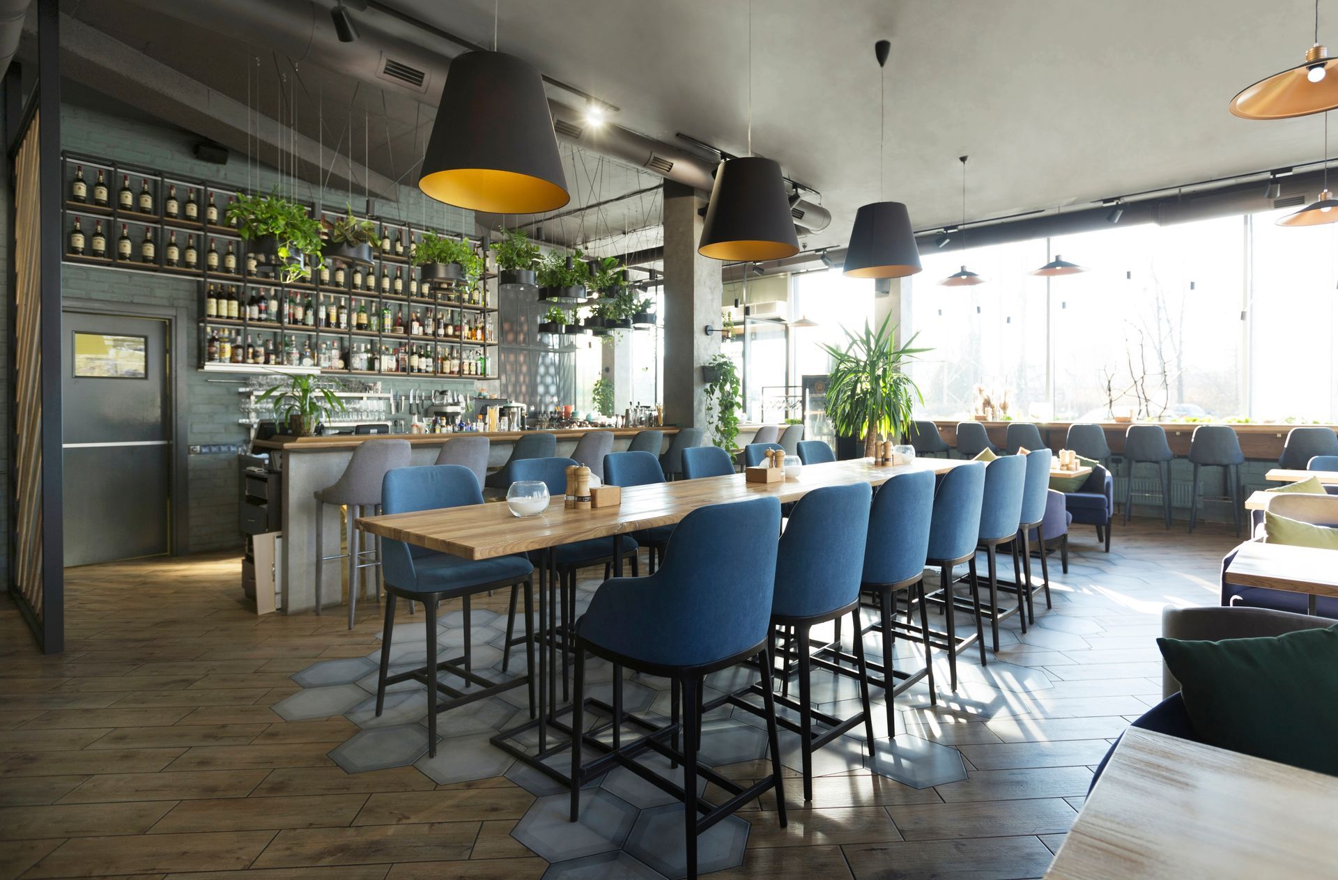 Modern restaurant interior with blue bar stools, wooden tables, hanging lights, and large windows