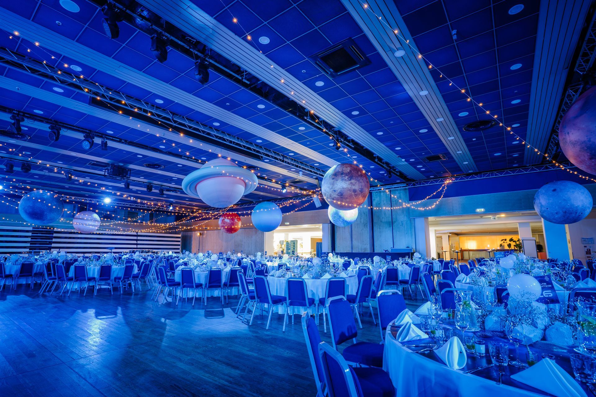Blue-lit banquet hall with round tables, chairs, and hanging globe decorations set for an event