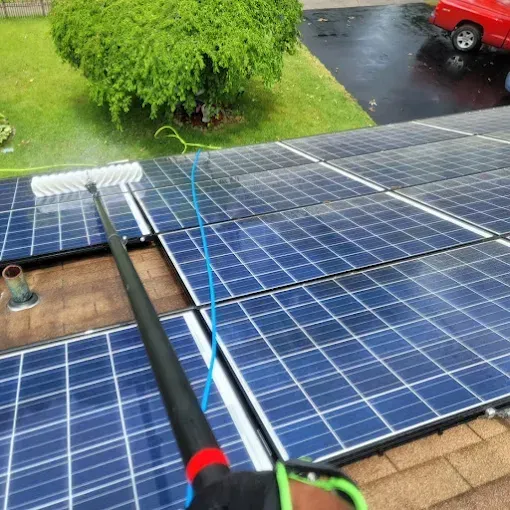 Pressure washing solar panels on a wet rooftop with a red car and lawn in the background