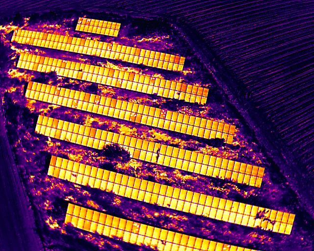 Person using a thermal camera to inspect solar panels on a rooftop. The camera is displaying a thermal image.
