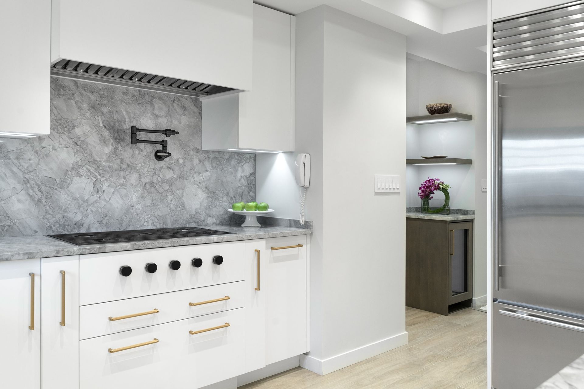 White kitchen with marble backsplash, range hood, gold hardware, and stainless refrigerator