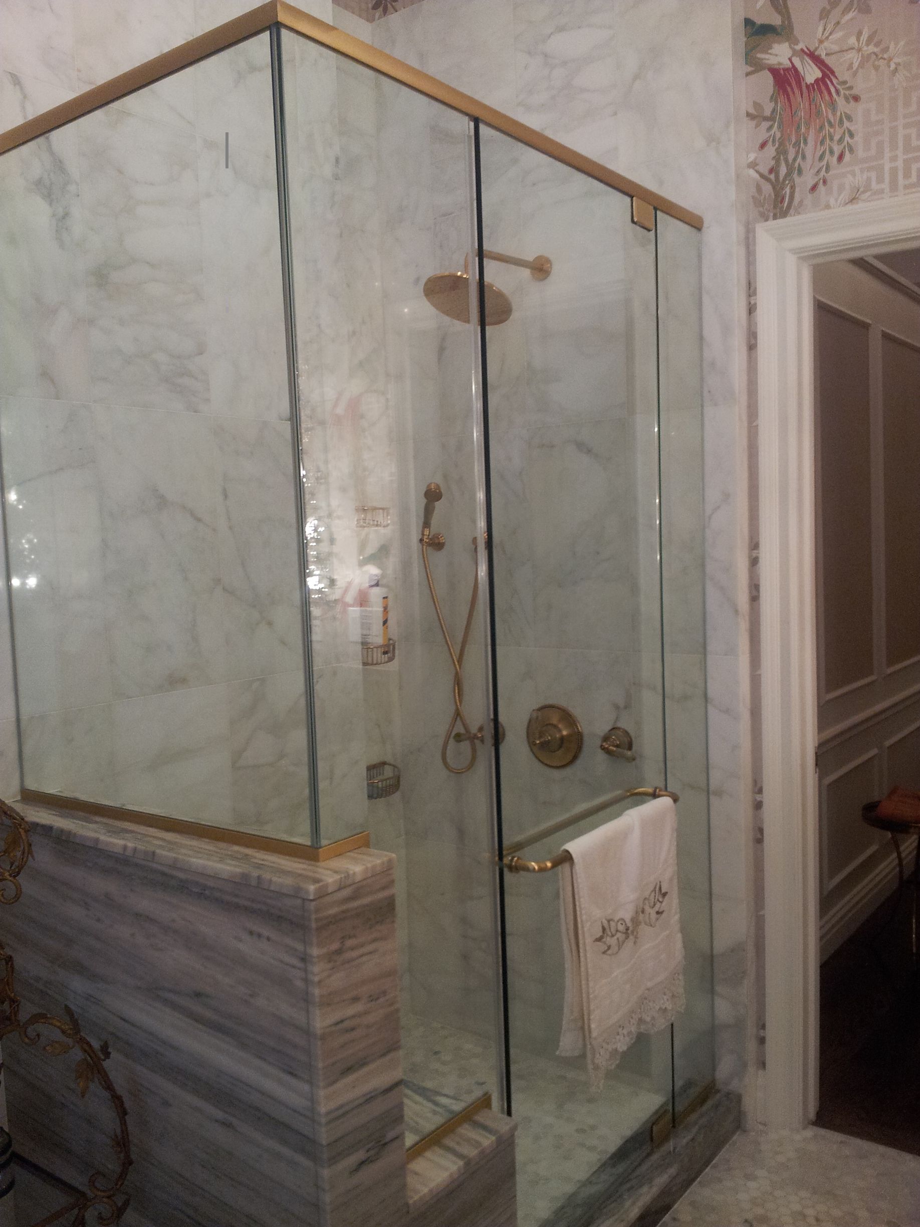 Marble bathroom shower with glass enclosure, brass trim, floral wallpaper, and brass fixtures