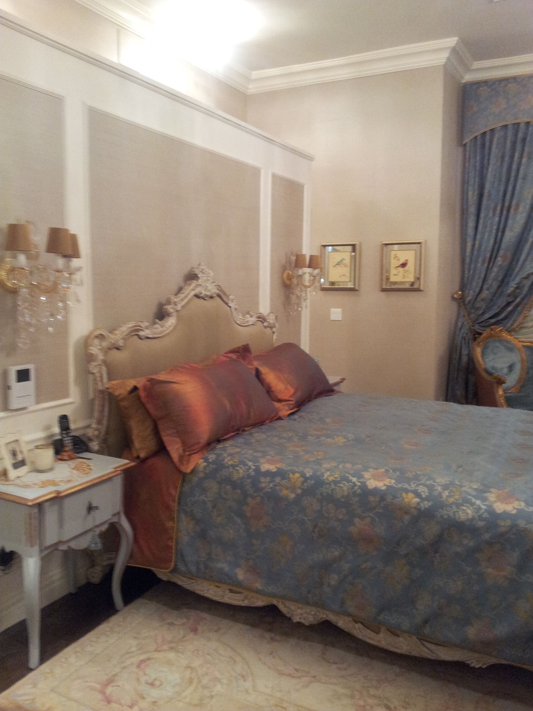 Traditional bedroom corner with upholstered headboard, framed artwork, crown molding, and blue drapery