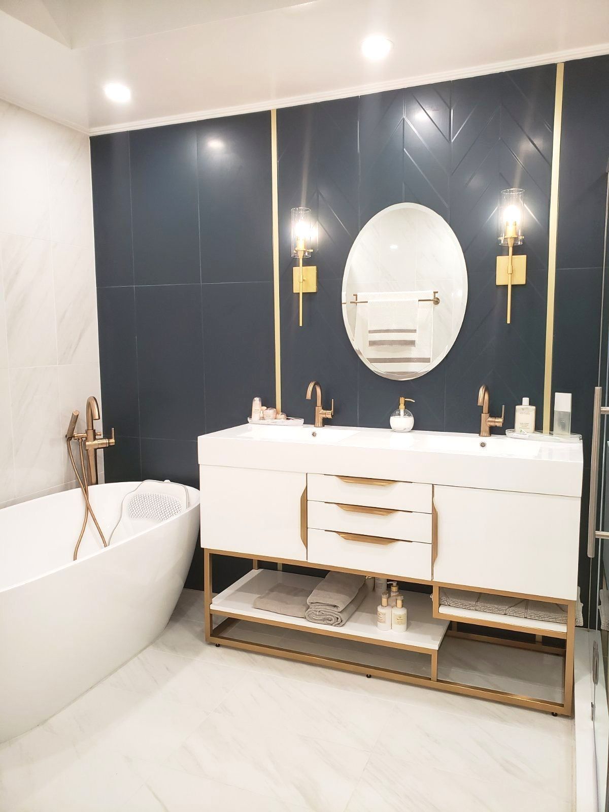 Modern bathroom vanity wall with navy tile, oval mirror, brass sconces, and marble accents