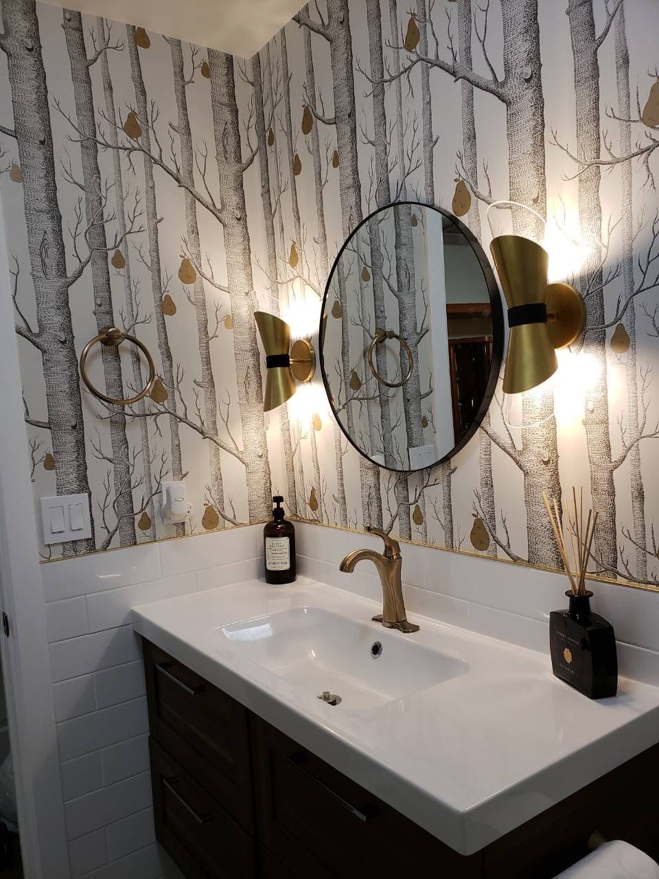 Bathroom wall with tree-pattern wallpaper, round mirror, and brass wall sconces