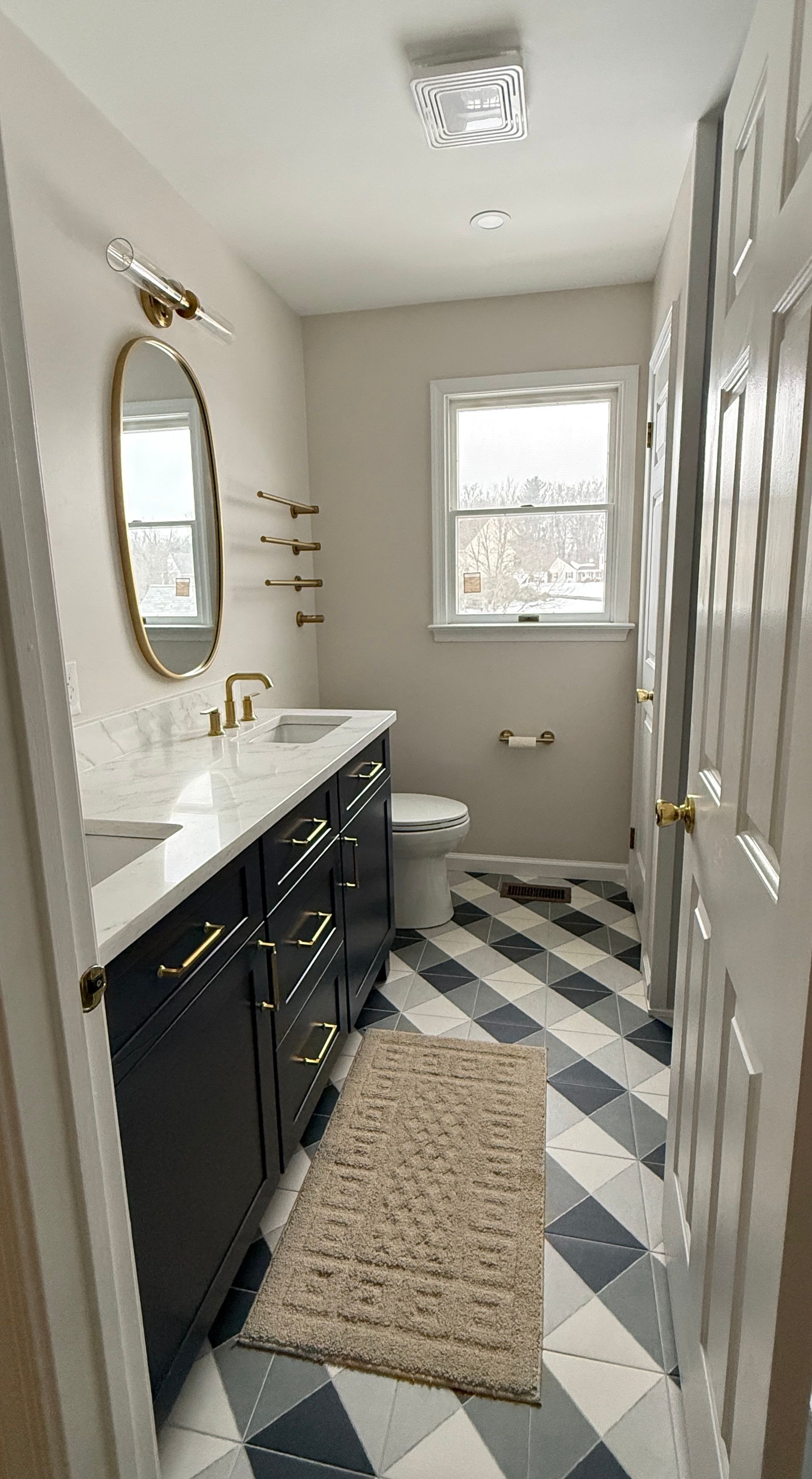 Neutral bathroom corner with arched mirror, brass wall sconce, navy double vanity, geometric floor tile, window, and ceiling vent